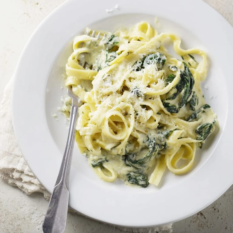 A close-up of Creamy Spinach Garlic Noodles, with steam rising from a creamy, golden sauce flecked with fresh spinach and garlic.